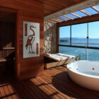 A modern bathroom features a blue heron artwork, large windows, a bathtub, and a scenic coastal view.