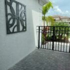 A decorative metal panel hangs on a white wall beside a gated patio area with pavers and tropical plants.