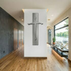 A contemporary crucifix hangs on a white wall in a modern hallway with wooden flooring and large windows.