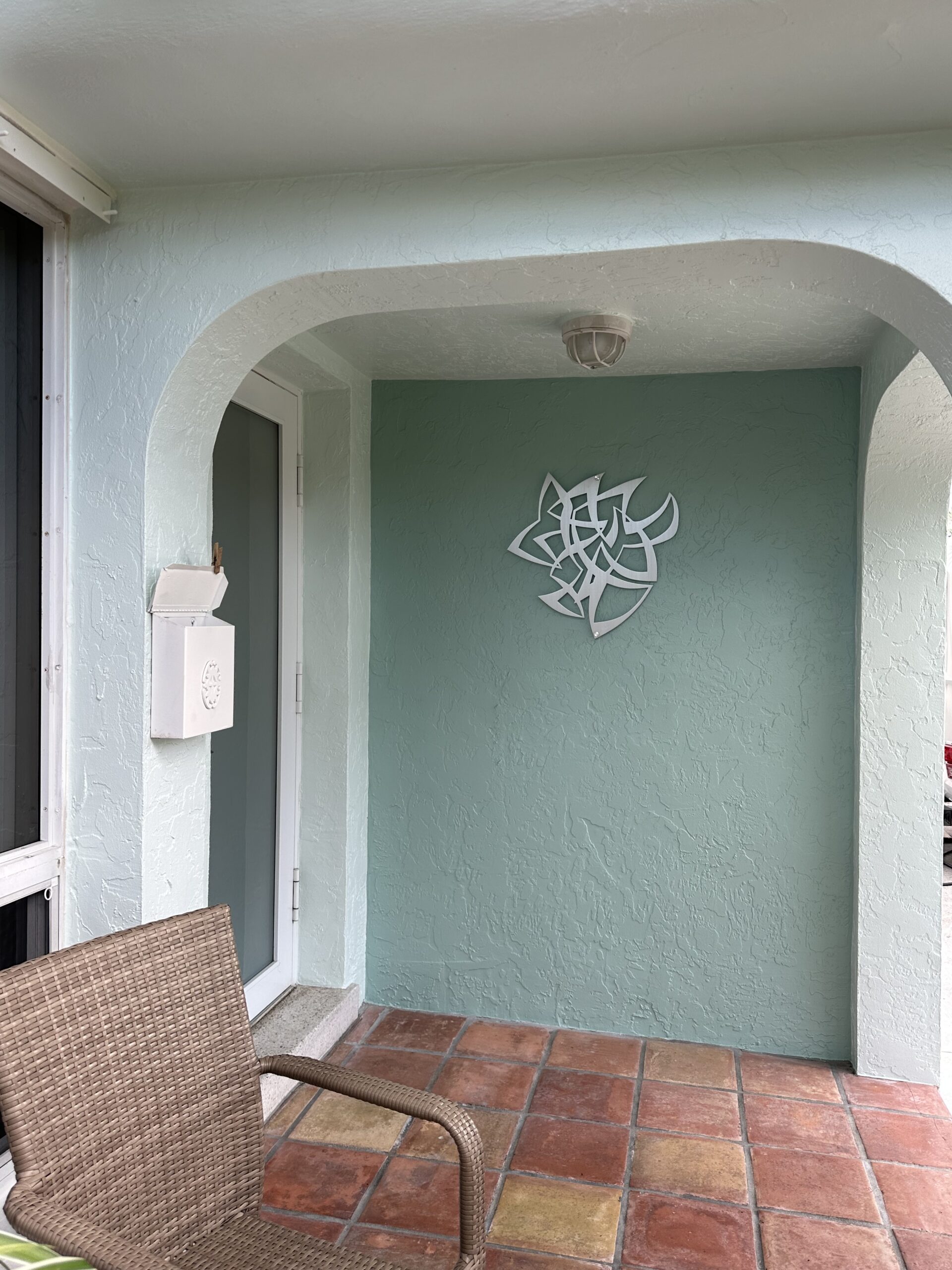 A small porch features a wicker chair, a light fixture, and a decorative wall piece on a mint green wall.