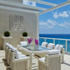 A modern outdoor dining area with a glass table, white chairs, and potted plants overlooking the ocean.