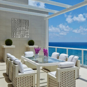 A modern outdoor dining area with a glass table, white chairs, and potted plants overlooking the ocean.