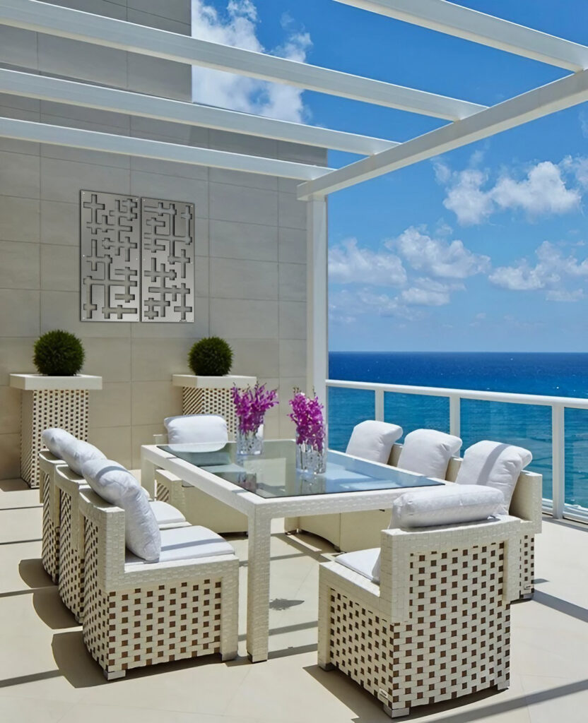 A modern outdoor dining area with a glass table, white chairs, and potted plants overlooking the ocean.