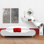 Two abstract metal artworks of nude figures hang on a white wall above a modern white sofa with red cushions.