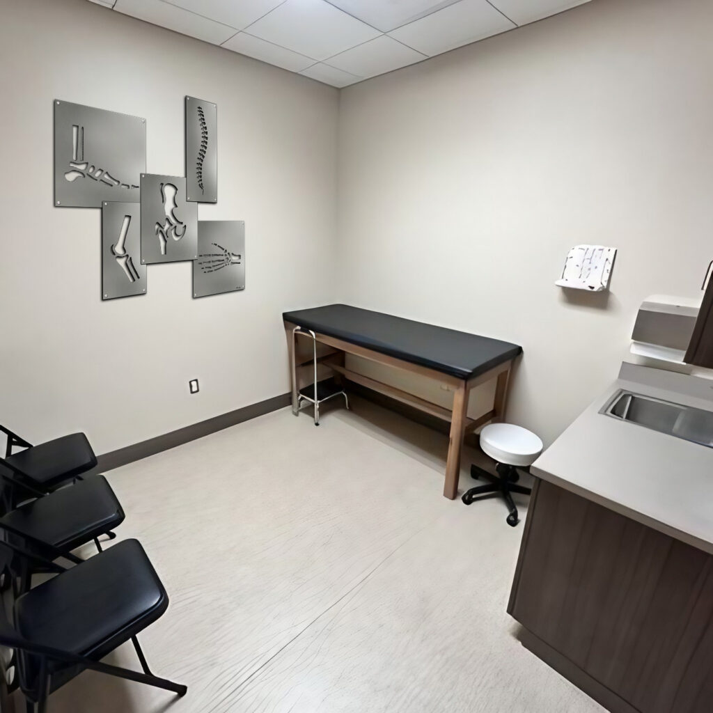 A medical examination room with a treatment table, chairs, and anatomical wall art.