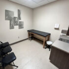 A medical examination room with a treatment table, chairs, and anatomical wall art.
