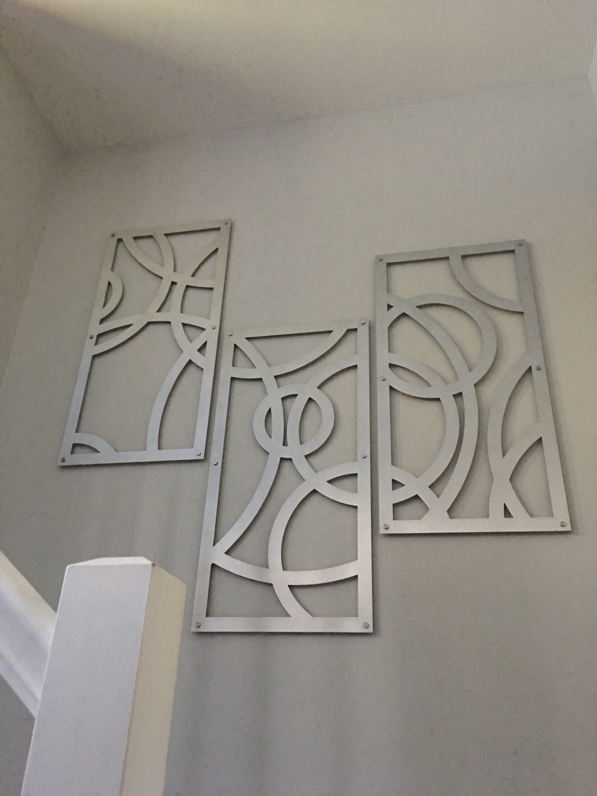 Three decorative metal panels with abstract designs hang on a light-colored wall near a staircase.