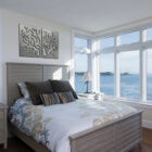 Bright bedroom with a bed, decorative pillows, and large windows overlooking the water. Wall art features tree designs.