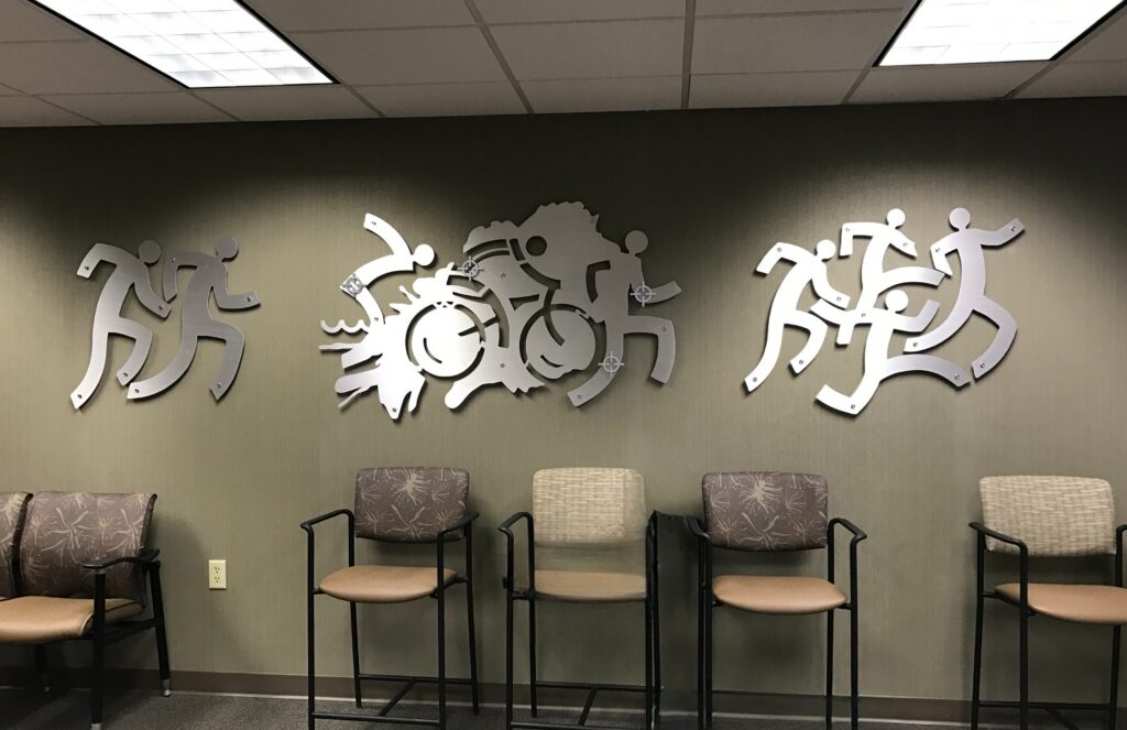 Three metal wall sculptures depict running figures and a cyclist, with empty chairs in the foreground.