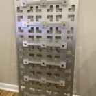 Metal panel with a geometric pattern of cutouts and decorative fasteners, mounted against a wall.
