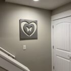 Framed art of two interlocking hearts with a butterfly in the center, mounted on a beige wall above a light switch near a white door. Cold Edge Gallery label at the bottom.
