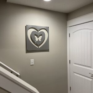Framed art of two interlocking hearts with a butterfly in the center, mounted on a beige wall above a light switch near a white door. Cold Edge Gallery label at the bottom.