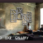 Two people seated in a modern art gallery with abstract metal wall art, concrete walls, and large windows. One is reading a book while the other looks at the artwork. Flowers are on a nearby shelf.