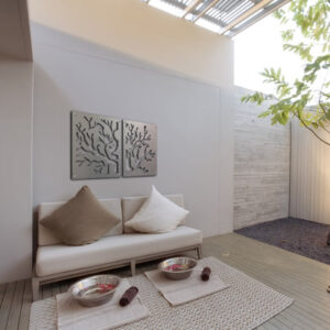 Outdoor minimalist seating area featuring a white couch with beige and brown pillows, metal tree art on the wall, and two metal bowls on a woven mat on the floor. A tree grows next to the space.