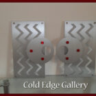 Two abstract metal art pieces with zigzag patterns and circular discs featuring red accents. Below, text reads "Cold Edge Gallery.