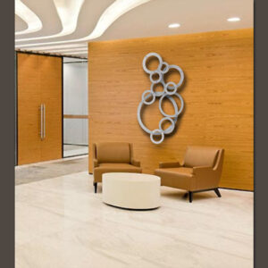 Lobby area with two brown leather chairs, a small round table, and a wooden accent wall featuring a circular custom metal artwork piece. A glass door allows light to stream in, and text on the wall reads "Cold Edge Gallery.