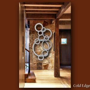 A custom wall-mounted metal sculpture of interlocking circles is displayed in a rustic interior with wooden beams and stone walls. The image includes the text "Cold Edge Gallery" in the bottom right corner.