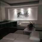 A modern living room with a sleek, minimalist design, featuring a custom metal wall art piece as the focal point. The artwork, composed of abstract, flowing shapes, is illuminated by subtle recessed lighting, creating soft shadows on the wall. The room is furnished with white leather sofas arranged in an L-shape, a glass coffee table with a potted plant, and clean, contemporary lines throughout the space. The overall ambiance is calm and sophisticated, emphasizing the elegance of the metal art installation.
