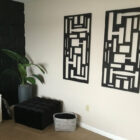 A room corner with two abstract black wall art pieces, a potted plant, a black ottoman, a woven basket, and a white dresser with a lamp.