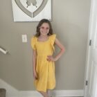 A young girl stands indoors, smiling, wearing a yellow dress with ruffled sleeves and brown high-heeled sandals. Behind her, there is a butterfly artwork in a heart frame on the wall.