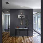 Art gallery interior with a modern cross sculpture on a dark grey wall, a slender black console table beneath it, and a plant. The walls have "Cold Edge Gallery" text vertically on the right side.
