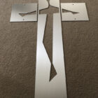 A silver metallic sheet with cutouts depicting a figure resembling a crucifixion, laid flat on a beige carpet.