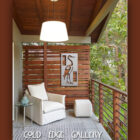 A cozy balcony with a white armchair, ottoman, and small round table. A wooden privacy screen displays art of a crane. The area is adorned with a patterned rug and lit by a hanging ceiling light.