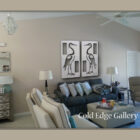 A contemporary living room featuring a gray sofa, white armchairs, and a coffee table. Two framed artworks depicting herons and labeled "Cold Edge Gallery" are displayed on the wall.