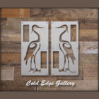 Two metallic panels featuring heron designs are mounted on a wooden wall and framed with a dark border, labeled "Cold Edge Gallery" at the bottom.