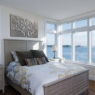 A well-lit bedroom with a large window overlooking a body of water. The bed has white bedding with floral patterns and multiple pillows. Two metal tree art pieces hang above the bed.