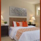 A bedroom with a wicker headboard features a bed with white and orange bedding. Above the bed is a three-piece metal wall art of trees. There are two nightstands with lamps on either side of the bed.