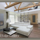 A modern living room with a white sectional sofa, wooden floors, exposed beams, and a minimalist style. The background features a loft area and a kitchen. Text reads: "Cold Edge Gallery.