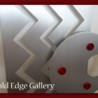 A geometric metal artwork featuring zigzag patterns and a circular element with red accents. The text "Cold Edge Gallery" is displayed in the lower-left corner.