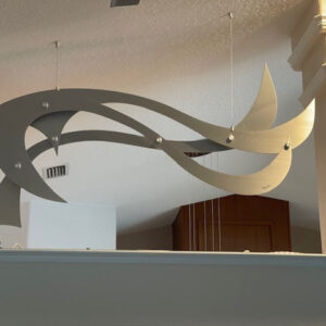 A modern metal sculpture with flowing, curved shapes hangs from the ceiling in a bright interior space.