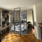 A room interior featuring a three-panel decorative metal divider with geometric patterns, partially separating a bedroom area. The panels hang from the ceiling, with the Cold Edge Gallery logo displayed at the bottom of the image. The room is decorated with posters on the walls and a wooden floor.