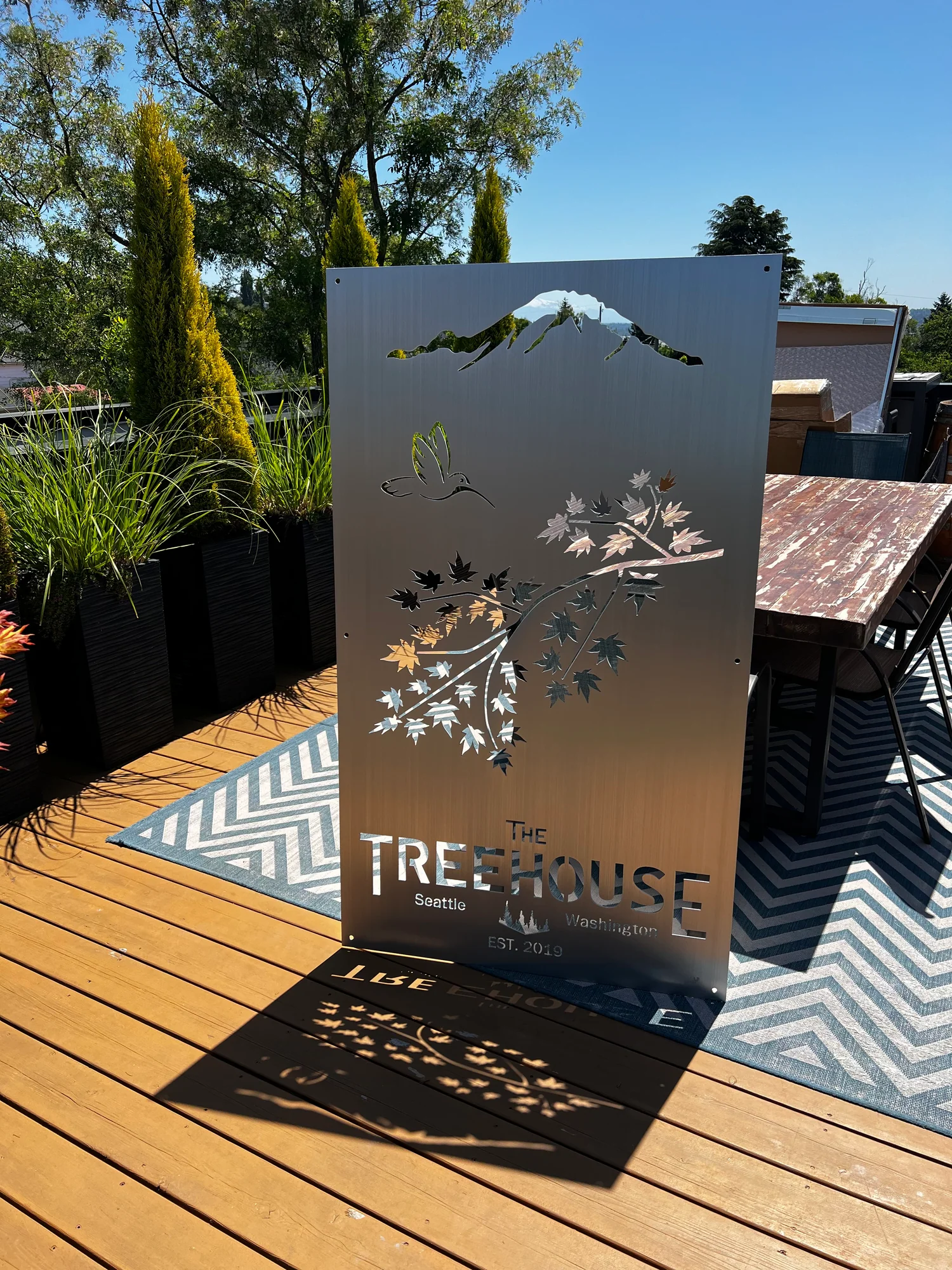 Metal sign for "The Treehouse" in Seattle, featuring a mountain, bird, and leaf designs, casting shadows on the deck.