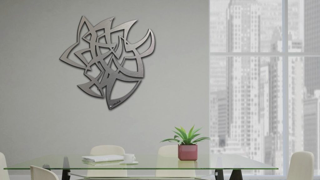 A modern metal wall art piece hangs above a glass table with a potted plant in a bright room.