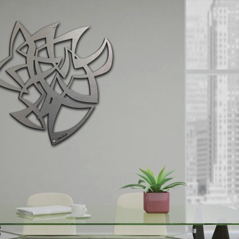 A modern metal wall art piece hangs above a glass table with a potted plant in a bright room.