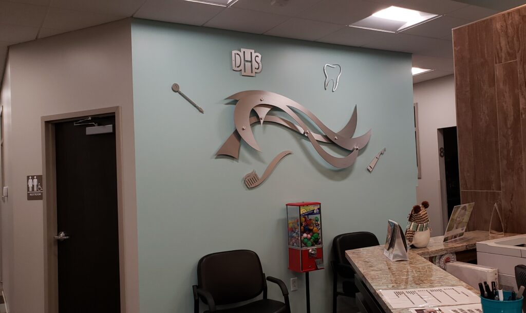 custom-sign-for-dentists-office-4 Metal dental-themed wall art with "DHS" logo, dental tools, and a gumball machine in a waiting area.