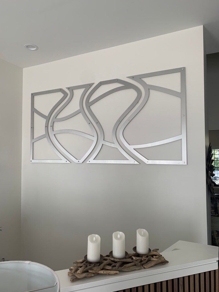 Metal wall art with abstract curves above a table featuring three white candles on driftwood.