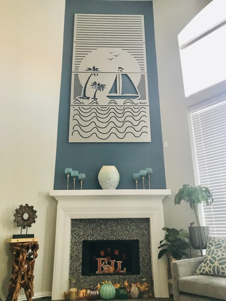Metal wall art features a sunset scene with a sailboat, palm trees, and waves, above a decorative fireplace.