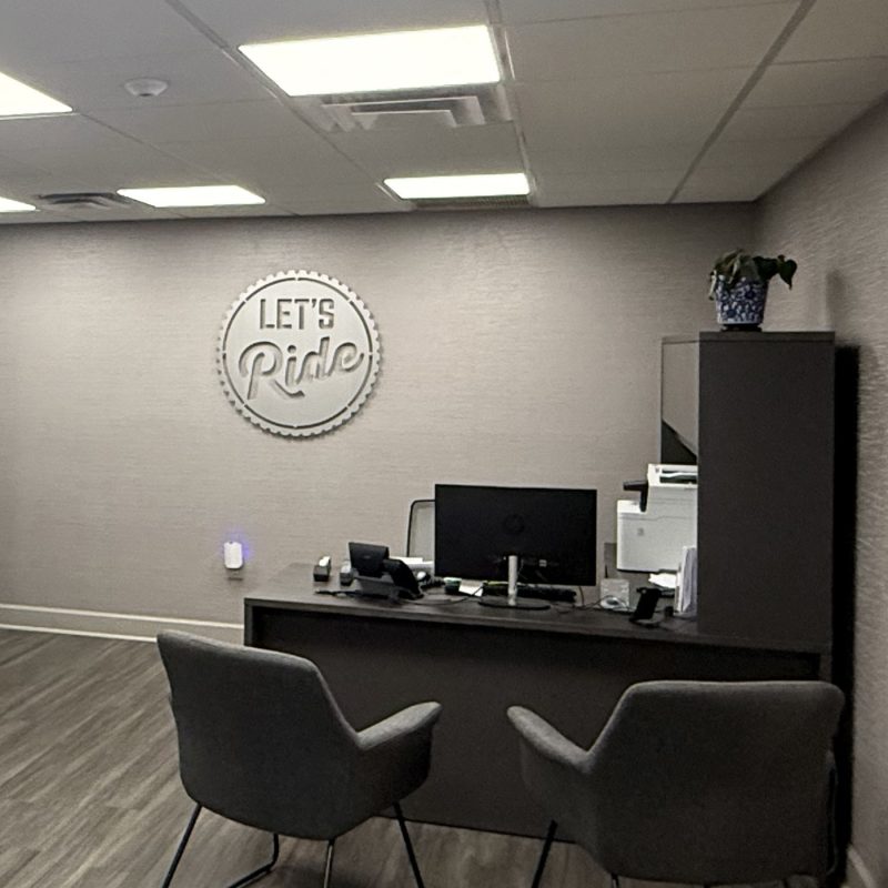 Office interior with a desk, two chairs, and a "LET'S Ride" sign on the wall. A plant sits on the desk.