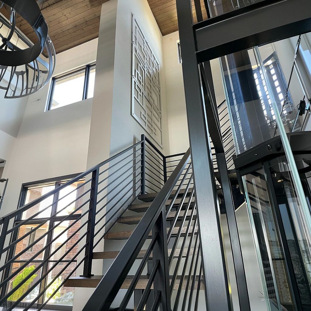 Modern staircase with metal railing, large windows, and a geometric wall art piece. Elevator visible nearby.