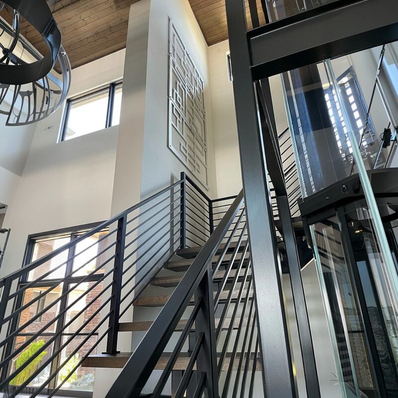 Modern staircase with metal railing, large windows, and a geometric wall art piece. Elevator visible nearby.