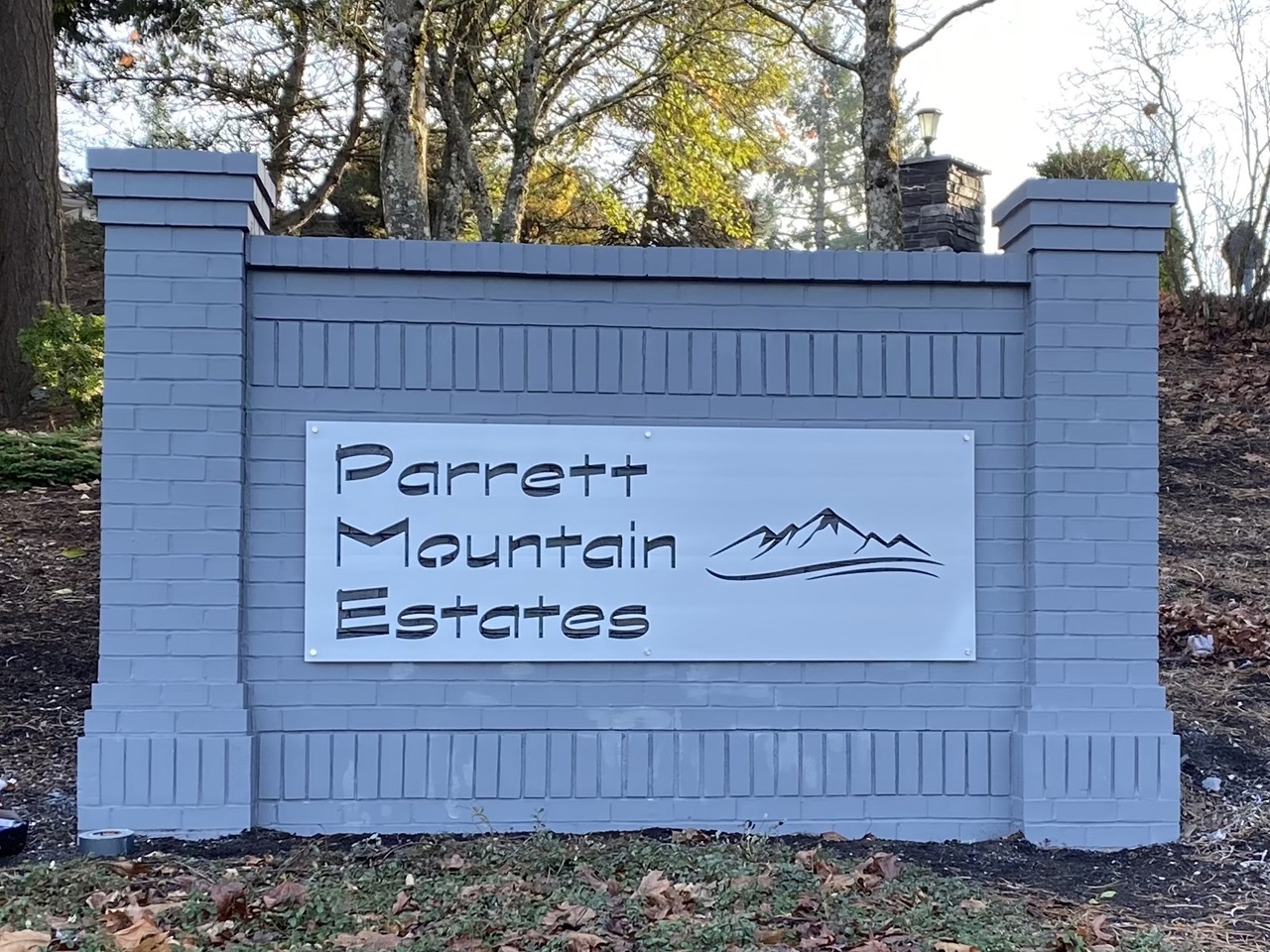 Sign for Parrett Mountain Estates displayed on a gray brick structure, surrounded by trees and fallen leaves.