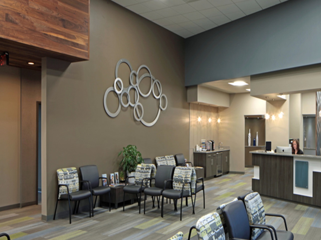 Modern waiting area with patterned chairs, a plant, and a reception desk. A person works at the desk.