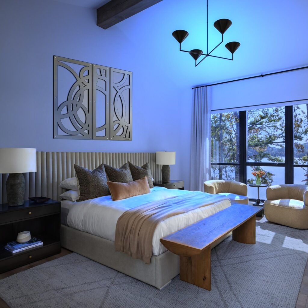 Modern bedroom with a large bed, decorative pillows, wooden bench, and two armchairs by a window.