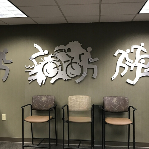 Three metal wall sculptures depict active figures running, cycling, and engaging in various sports. Empty chairs are visible below.